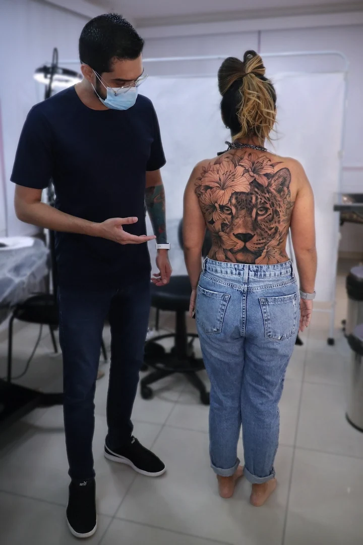 Lucas Franca ao lado de uma cliente com tatuagem realista de onça e flores nas costas